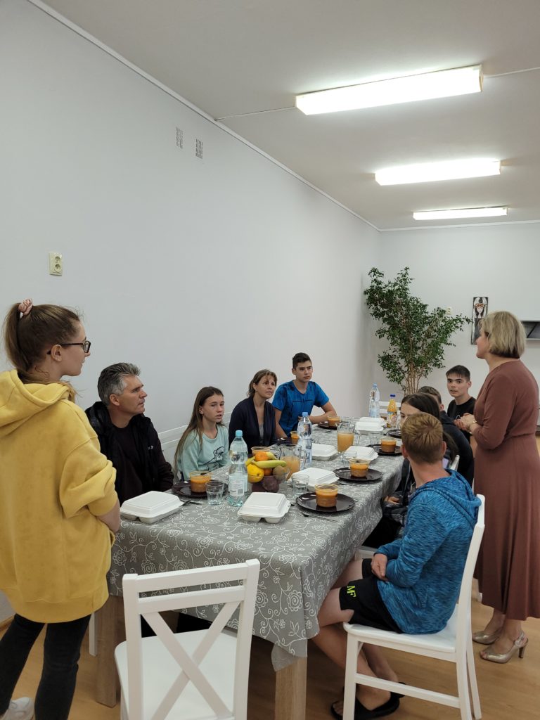 Zdjęcie rodziny zastępczej z Ukrainy siedzącej przy stole oraz Pani Dyrektor Małgorzata Malczuk stojąca obok stołu