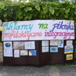 Baner powitalny z tekstem Witamy na pikniku profilaktyczno integracyjnym. Po banerem prace plastyczne dzieci.